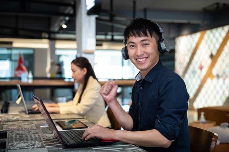 Happy young Asian man listening music with wireless headphones and working in co-working office. He cheering with excitement and looking at camera.