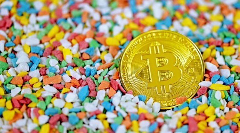 Graphic shows a gold Bitcoin coin laying on the background full of colorfull stones