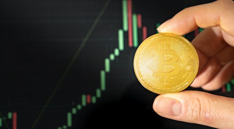 Person holds BTC token in fingers with a candle chart in the background