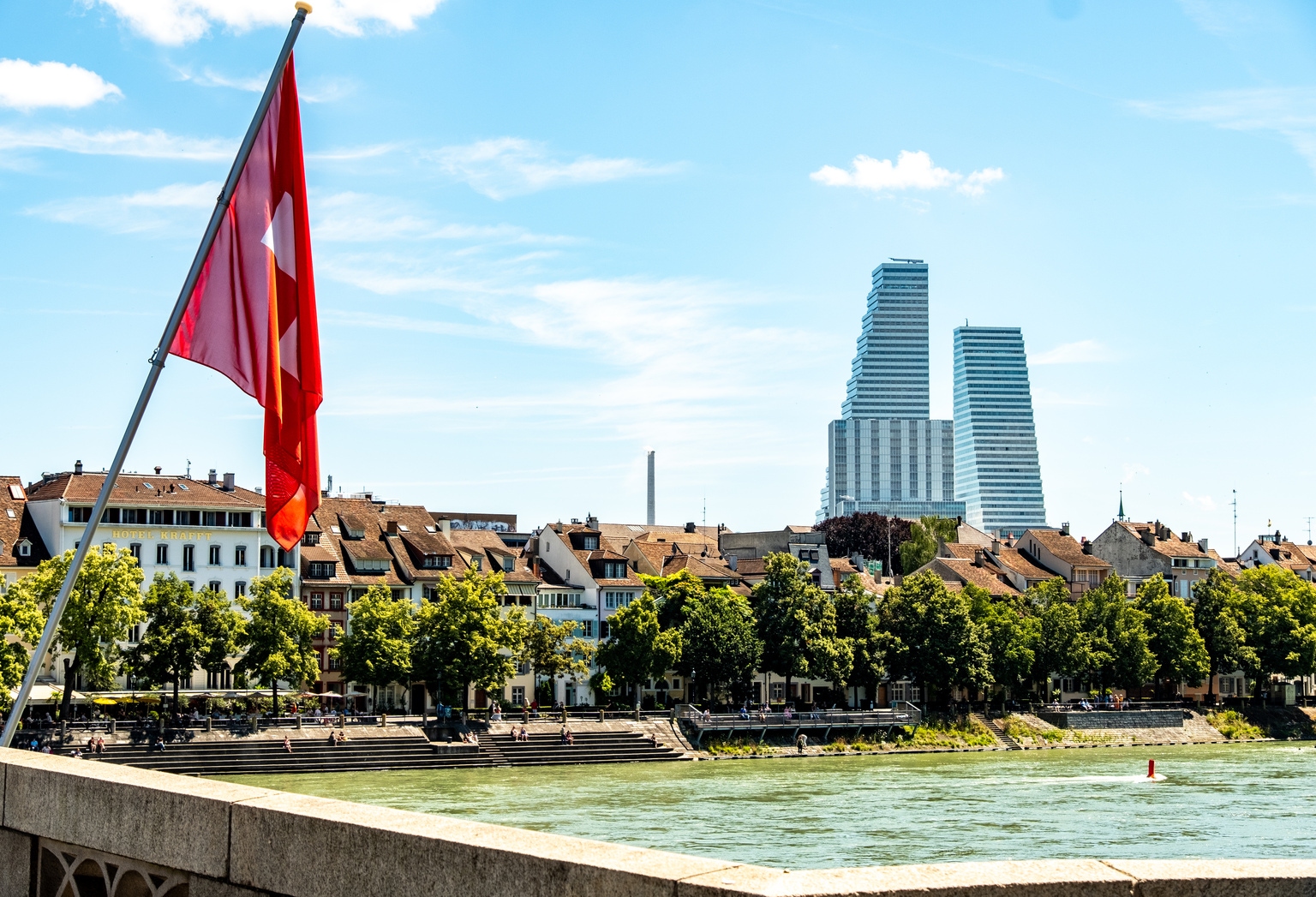Der Rhein in Basel mit einer Schweizer Flagge im Vordergrund und den Roche Towers im Hintergrund