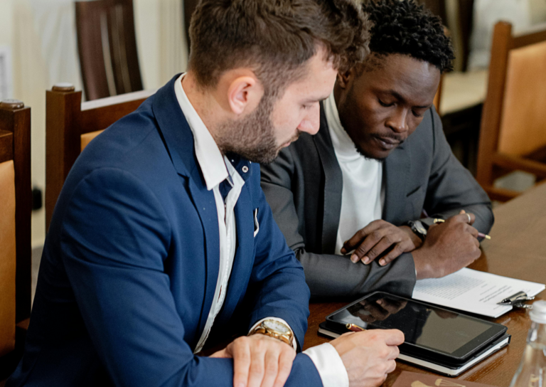 Two businessmen in suits review and sign documents at a wooden table with a tablet and papers nearby.