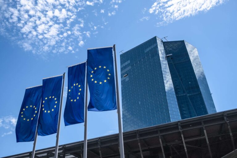 Row of flagpoles with European Union flags (blue with yellow stars) in front of a modern glass building against a clear blue sky.