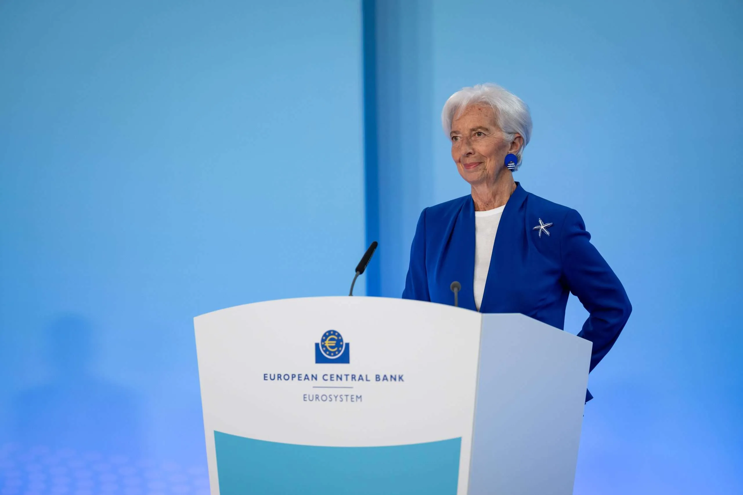 Senior woman at a white podium with ECB/European Central Bank branding, speaking in a blue backdrop, wearing a blue blazer and white blouse.